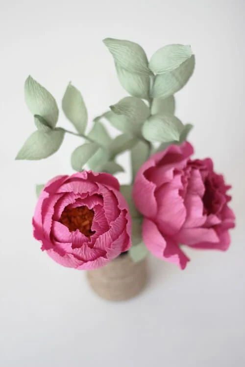 pair of deep pink crepe paper peonies with green foliage in vase