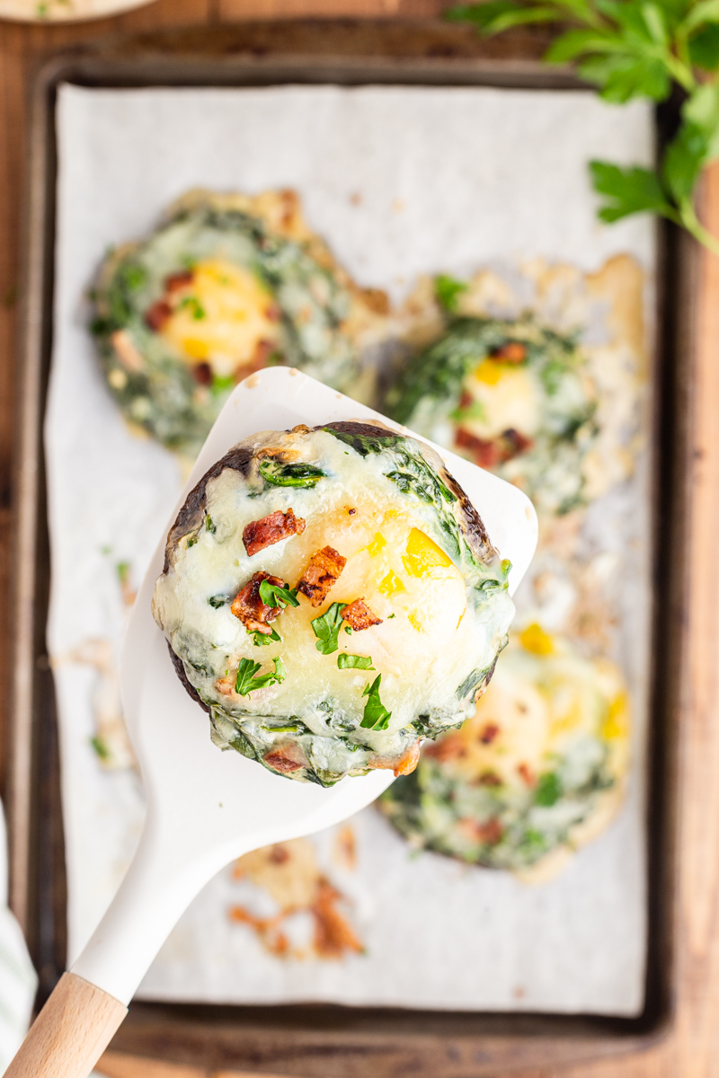 Overhead photo of Keto Spinach and Egg Stuffed Mushrooms on a baking sheet with one on a spatula closer to the camera.