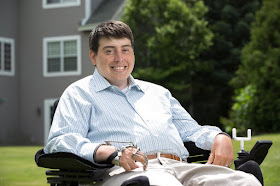 Photo of Jimmy Anderson, a young, half Hispanic, half white man with short brown hair. He is sitting in a power chair with a long-sleeved blue and white striped shirt and khaki pants. Behind him is a green lawn, a home and tree. He is smiling at the camera.