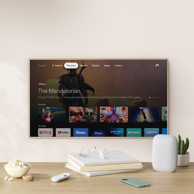 A TV mounted on the wall shows the Google TV experience. A counter sits in front of it with a Nest Audio, Chromecast remote, Pixel phone, stack of books, bowl of popcorn and small cactus plant.