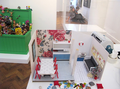 Dolls' house miniature scene, being built on  a table in a gallery during install. In the background, two gallery staff discuss placement of signage on the wall.