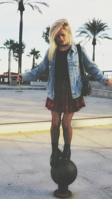 Estilo grunge con camiseta negra, falda de tablas tartán, medias de red grande, botas estilo militar y cazadora vaquera