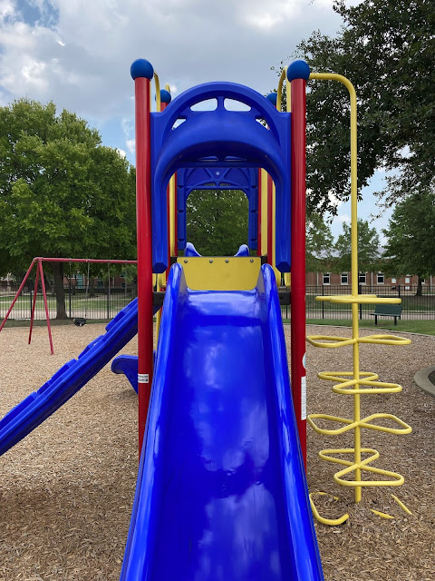 A photo of playground equipment that is red, blue, and yellow. A blue slide and the landing behind it are in the center of the photo, with a blue climbing wall on the left-hand side and some yellow climbing bars on the right-hand side. A large tree, a swing set, and a building are visible in the background.