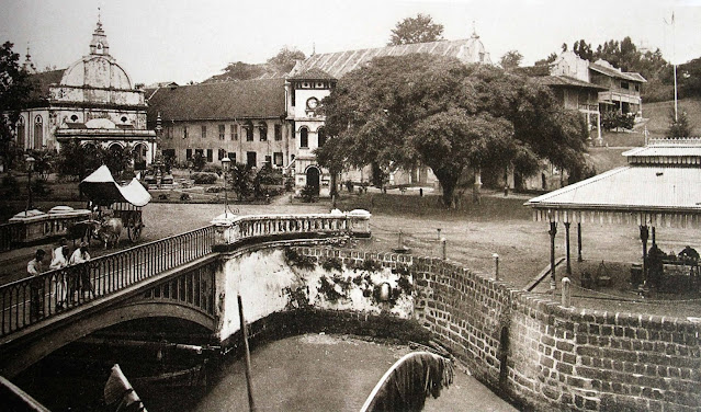 A Melaka bullock carts head over the Melaka bridge from the Town Square with Christ Church (left), the Clock Tower (center) and the Stadthuys behind, c. 1906.