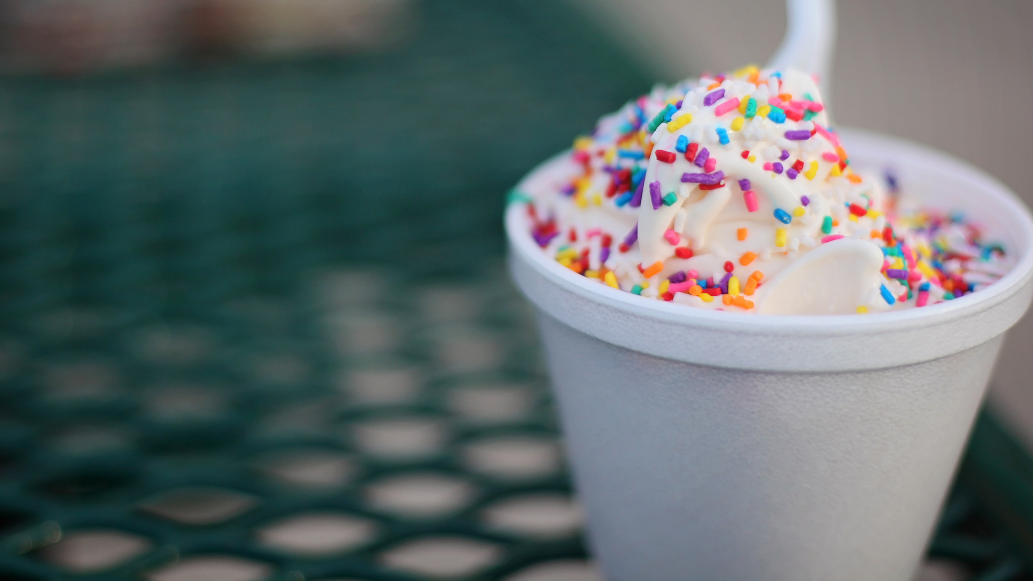 ice cream in a cup on a wire table. There are sprinkles on the ice cream and a plastic spoon in it.