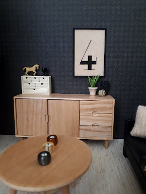 One-twelfth scale modern miniature lounge in cream and black, with a framed printed number four on the wall above a scandinavian sideboard. On the coffee table in the foreground are a row of three candles, under the picture is a plant and two vases, and on the other end of the sideboard is a set of mini drawers and two ornaments