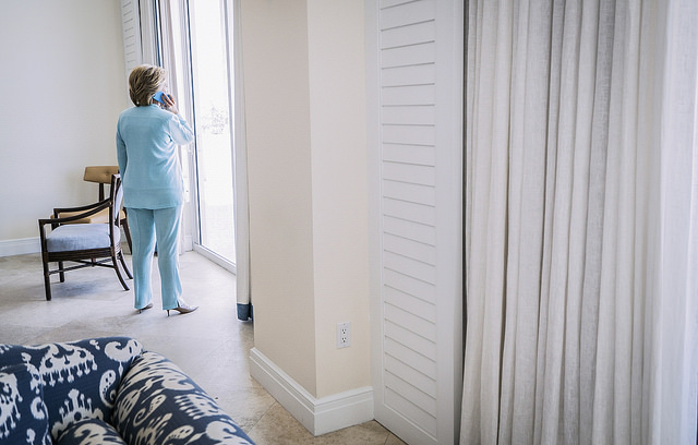 image of Hillary Clinton standing in what looks to be a hotel room with her back to the camera, speaking on a cell phone while she gazes out a window