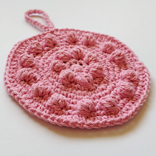 Pink round cotton crochet dishcloth with bobbles resting on white background.