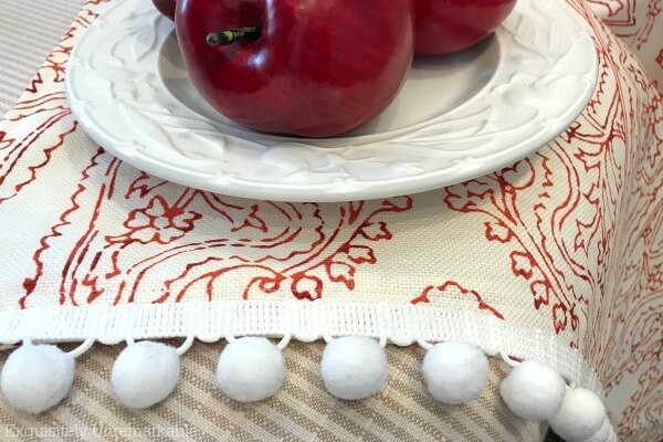 Red and white fabric with white ball fringe draped over chair with a white plate and apples on top
