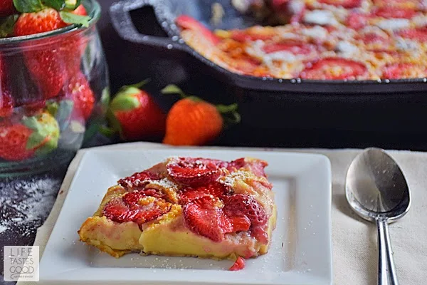 A slice of strawberry clafoutis on a white plate with a spook along side. A fresh bowl of strawberries in the background with the skillet of strawberry clafoutis