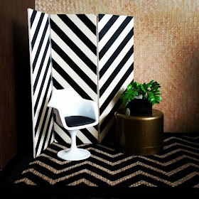 One-twelfth scale modern miniature scene including a plywood screen with black stripes, a hessian rug with a black chevron pattern, a white tulip chair with a black seat and a bronze side table with a potted fern sitting on it.