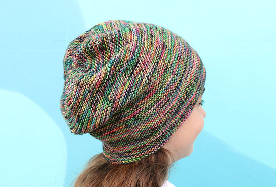 Back view of girl wearing colorful slouchy hand knit Rikke hat with a bright blue background.