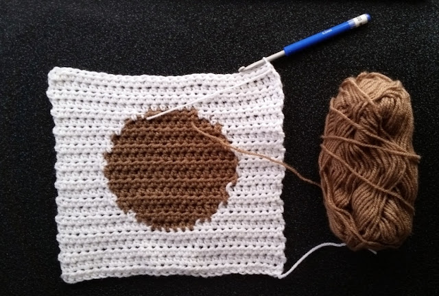 The reverse side of a white square with a tan circle in the middle. The square is worked in rows using intarsia technique for colour changes. A crochet hook with a blue handle is still inserted into the last stitch of the top row, on the top right-hand corner of the square. A skein of tan yarn is still joined to the circle and sits to the right of the square.