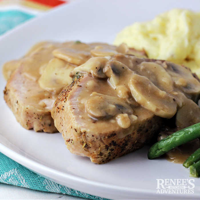 2 slices of pork ribeye roast with mushroom gravy on top on a white plate with mashed potatoes and green beans