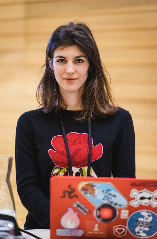Image shows  Katerina Skroumpelou looking straight ahead at the camera. She is seated behind a laptop with a red cover covered in stickers