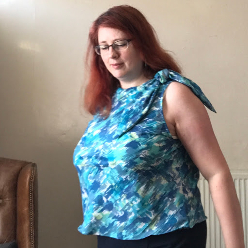 A redheaded woman in a top that ties at one shoulder. The top is in various shade of blue, plus a splash of spring green.