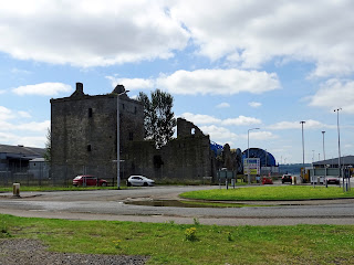 A photo of Rosyth Castle with huge blue circular structures behind it on which cables to be laid on the sea bed are rolled up into as well as views of other buildings in the dockyard.  Photo by Kevin Nosferatu for the Skulferatu Project