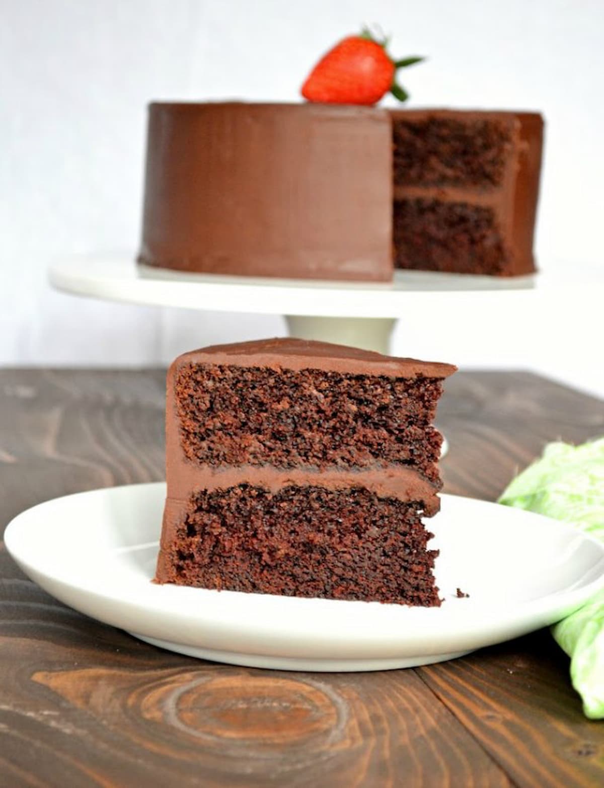 A slice of Chocolate Cake with Ganache Frosting on a white plate with the whole cake in the background on a white cake stand.