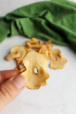 Make your own apple chips with very little effort. Just bake them in your oven and they are ready to be added to salads, used as garnishes for desserts or just be snacked on! hand holding up a homemade apple chip with more apple chips in backgound