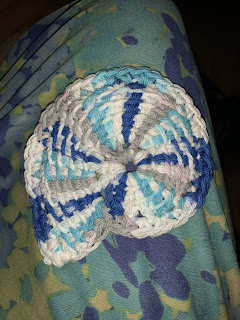 A Tunisian crochet seashell in the shape of an ammonite, in the white yarn with splashes of gray, aqua, and blue forming rays from the center out.