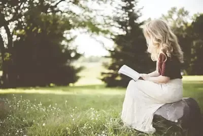 A woman sitting on a rock reading an inspirational and motivational book in a grassy field.