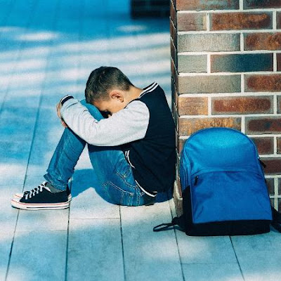 #pracegover – um menino de tênis, calça jeans e moleton, sentado no chão e encostado em uma coluna de tijolo, cabeça entre os joelhos, ao lado sua mochila.