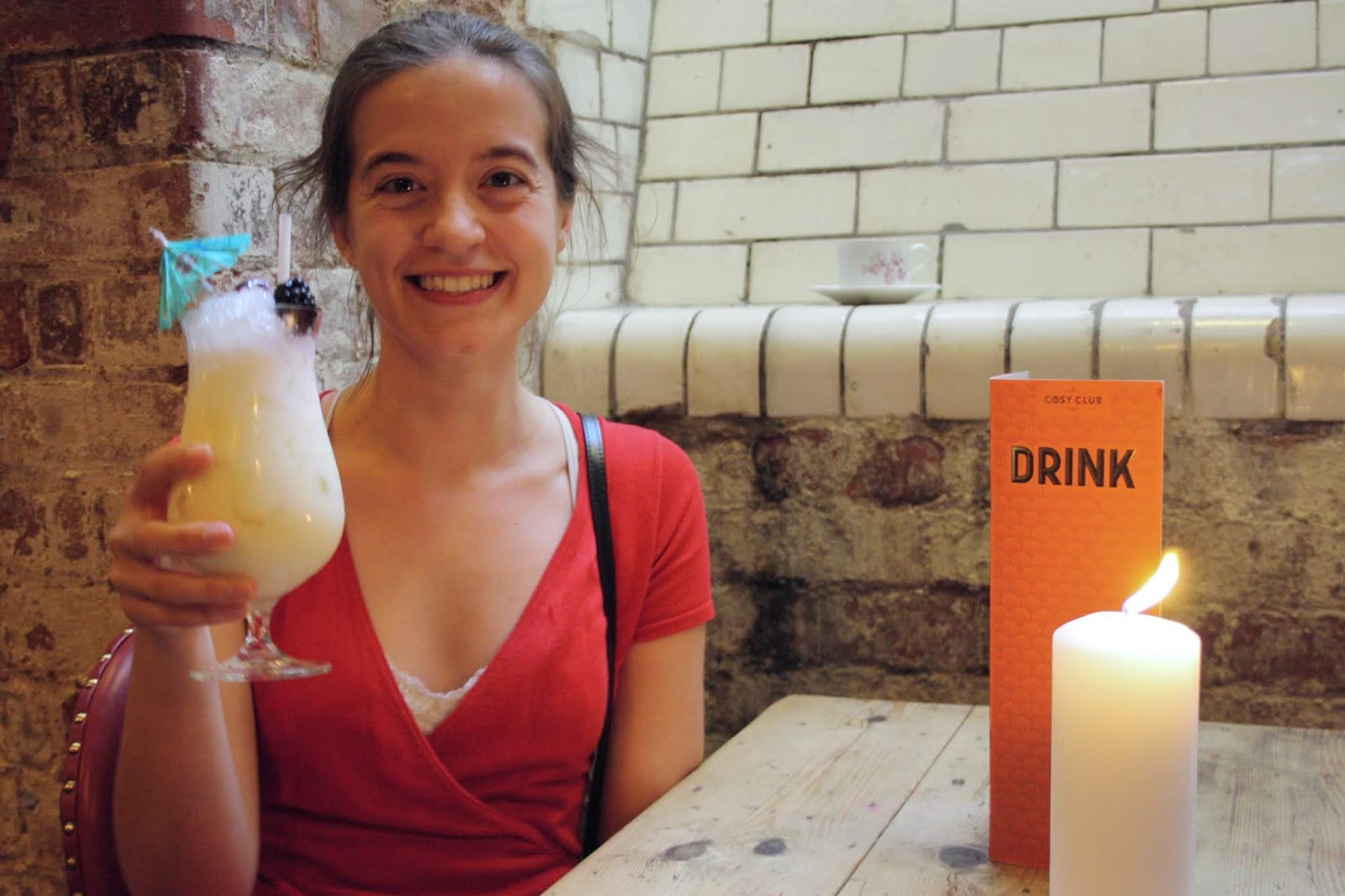 Abbey, wearing a red dress, holds up a Pina colada cocktail, which is decorated with fruit and a cocktail umbrella