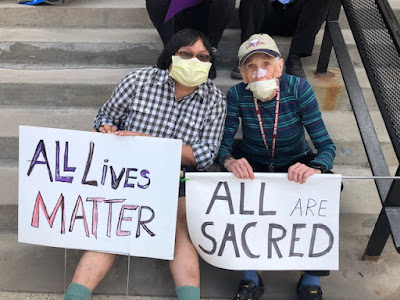 Two people in masks holding signs: one darker skinned and taller,the other white and a little stooped 