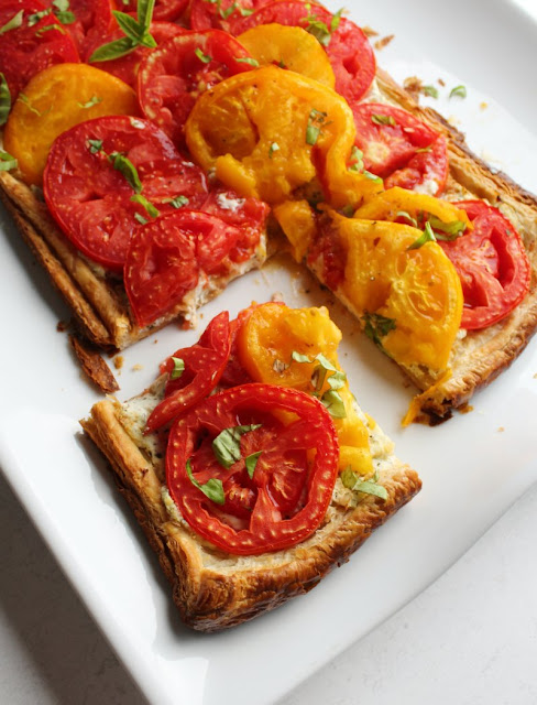 Fresh slices of vine ripened tomatoes, a basil and ricotta cheese filling and golden puff pastry crust come together to make this delicious flaky tomato tart. It is a perfect summer appetizer, lunch or light dinner when paired with a salad. corner slice of flaky ricotta and tomato tart with yellow and red tomatoes and fresh basil on top