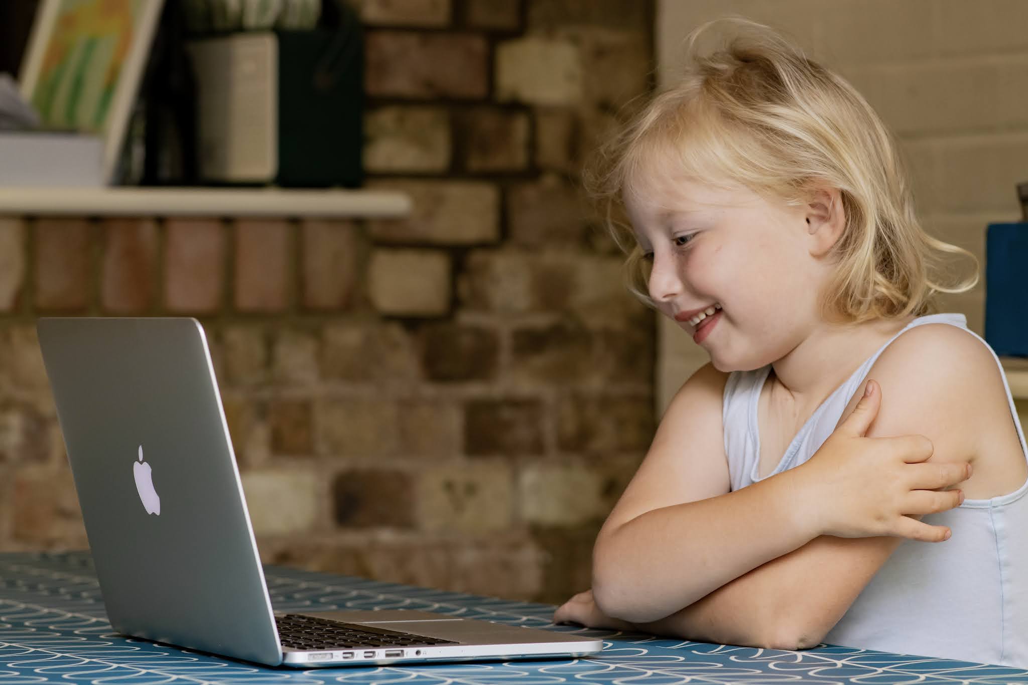 A EYFS Key stage 1 primary age girl smiling at a laptop during an online tutoring session