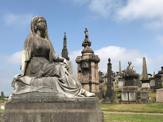 View over gravestones and memorials - the photo shows in the foreground the statue of a sitting woman looking up to the sky.  Part of her face has been chipped off.  Photo by Kevin Nosferatu for The Skulferatu Project.