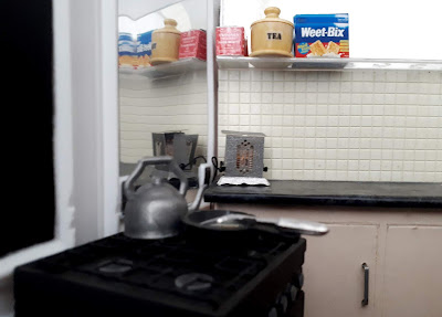 1/12 scale miniature kitchen, On the stove is a kettle and a frying pan and fish slice. On the bench is an old-fashioned toaster, and on the shelf above it is a tin of Twinings tea, a tea jar and a box of Weetbix. 