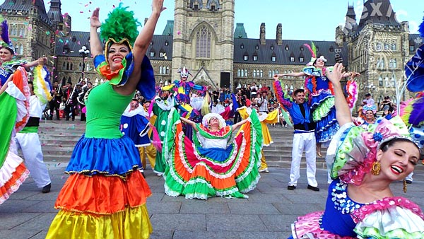 several women and a couple of men are dancing in a plaza in front of an English-looking church. The women wear brightly-colored, layered dresses.