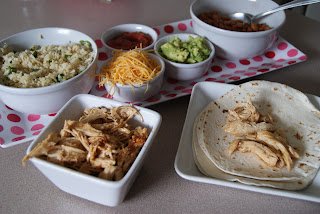 A flour tortilla topped with shredded chicken served on a plate, along side of bowls filled with cilantro lime rice, shredded cheese, guacamole, black beans, shredded chicken and diced tomatoes.