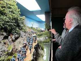 Two men in front of a diorama of 19th-century soldiers marching through the bush.