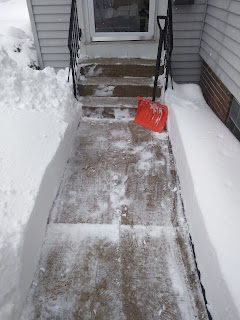 Orange snow shovel standing against concrete stairs to house with shoveled sidewalk and deep snow
