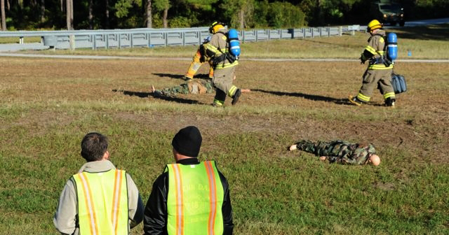 Two men oversee a training exercise - there are two people in protective suits walking across a field.