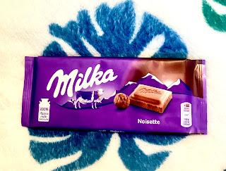 A rectangular purple chocolate bar with Milka in white font with a picture of a brown square chocolate piece next to a light brown oval hazelnut with Noisette in small white font written below on a bright background