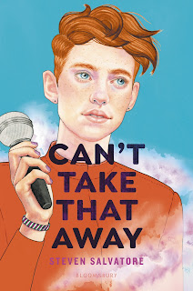 A white qenderqueer person with bright red/orange hair wearing an orange shirt is holding a microphone in their left hand. Their wrist has a green, white, and purple striped bracelet and their nails have purple polish.