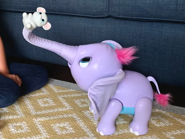 A purple, pink and blue elephant toy with it's trunk in the air holding a toy mouse