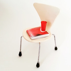 Modern dolls' house miniature white Arne Jacobsen series 7 chair, with a red book on Arne Jacobsen and glass on the seat.
