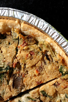 quiche in a tin pan with sundried tomato pieces, spinach, artichokes, and mushroom slices.