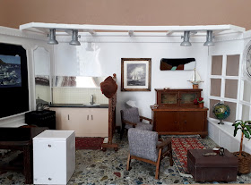 1/12 scale modern miniature scene of a single man's flat, with a simple kitchen on the left with a large vintage table and captain's chair, and a living room on the right with two mid-century modern armchairs, an antigue cupboard, battered sailor's chest and afghan rug.
