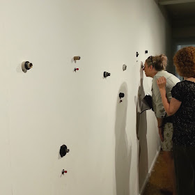 A woman peering into one of several small holes in an art gallery wall, while another woman looks on.