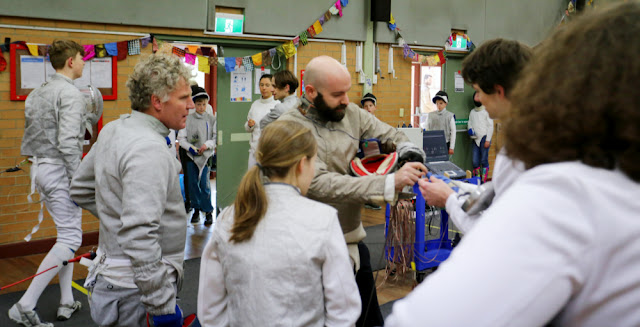 A group of fencers gather as Coach Joe Raciborski inspects a piece of equipment. They stand in a semicircle, dressed to fence foil, epee and sabre. In the background other fencers wait to commence a bout on one of the two pistes behind the group. There is bunting! Heaps of bunting! Hung up along the walls, and whaddaya know, there's that legend Marina Carrier taking time out from her Olympic training schedule to come and fence with the club.