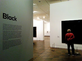 Entrance to an exhibition with the name of it ('Black') on the lefthand wall, with explanation text below. On the right a man in a red shirt is looking at an all-black art work.