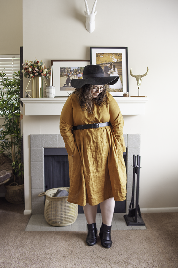 An outfit consisting of an oversized black floppy hat, a golden yellow collared button down linen dress, and black heeled Chelsea boots.