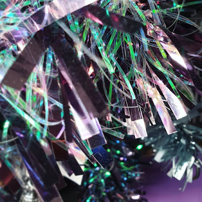 Iridescent tinsel Iridiscent green and purple tinsel with almost transparent fronds, against the background of a dark purple wall. The image is very busy, and the tinsel takes up most of the image