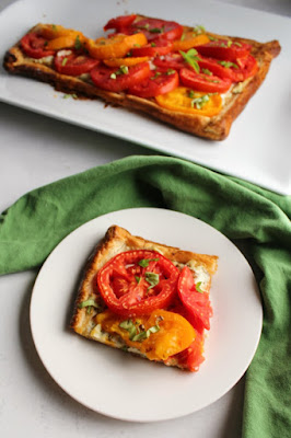 Fresh slices of vine ripened tomatoes, a basil and ricotta cheese filling and golden puff pastry crust come together to make this delicious flaky tomato tart. It is a perfect summer appetizer, lunch or light dinner when paired with a salad. First slice of tomato tart served on plate with remaining tart in background