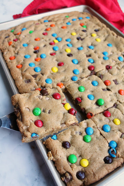Chewy brown sugary blondies loaded with chocolate chunks, caramel undertones and topped with M&Ms. This big batch of bars is perfect for a party, carry-in or just because. first piece of chewy salted caramel M&M blondies being lifted out of sheet pan with spatula